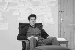 Black and white photo. Colin Morgan is sat in the leather arm chair. He's wearing the baseball style jacket and chino trousers. He's holding a mug and resting it on his lap, he looks still, not giving much away. In the background plastered across the walls are lots of bits of A4 paper with writing on. It looks like a time line of events but we can't see the writing.