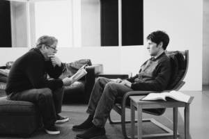 Black and white photo. Roger Allam and Colin Morgan face each other. Roger is sat on a cushioned foot rest from a sofa set and is leaning forward resting his elbows on his knees. He's holding a script and has glasses on the end of his nose, and looks over them intently at Colin. Colin is sat on the leather arm chair. He is sat back and looks on edge. In the surrounding room we see what makes up a living room set with a leather sofa behind them and a couple of wooden side tables.