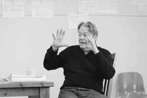 Roger Allam sat at a wooden table wearing a black v neck jumper and jeans. He's holding both hands out wide in front of his face as if indicating the size of something. His eyes are wide.
