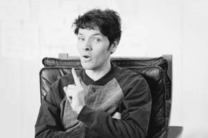 Black and white photo of a man (Colin Morgan) in his 30's. He's sat in a dark leather arm chair. He is wearing a baseball style jacket and has one arm folded across his body, and the other is raised with his finger in his point. His face is stern like he might explaining something.