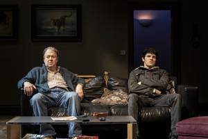 A drab living room. In low light we see Roger Allam and Colin Morgan sat apart on a leather slouchy black sofa. Roger is slouched back and his lip is curled like he's angry. Colin is sat back with his arms crossed, looking furious. In the dark background there's a doorframe leading out into a hallway, and on the back walls are large framed paintings, one of a tiger.