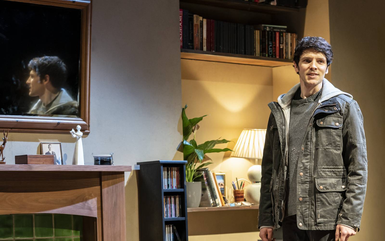 Colin Morgan as Bernard stands in a modern living room, in front of his reflection in the mirror