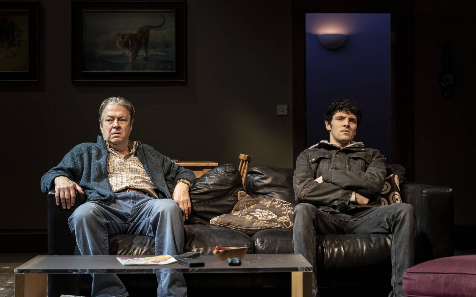 Roger Allam (father) and Colin Morgan (son) sit on a sofa, leaving space between them. They look unhappy