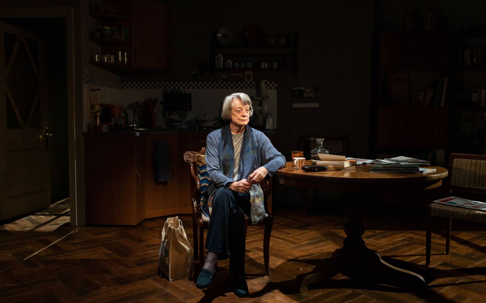 An older woman sits alone in a darkened kitchen, her elbow leaning against a table