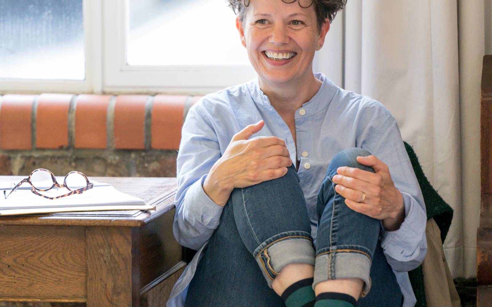 Sally Cookson sits on a chair, smiling, with her knees hugged up to her chest