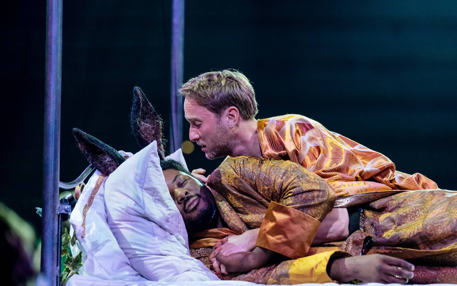 Hammed Animashaun (Bottom) sleeps in a bed, as Oliver Chris (Oberon) leans lustily over him