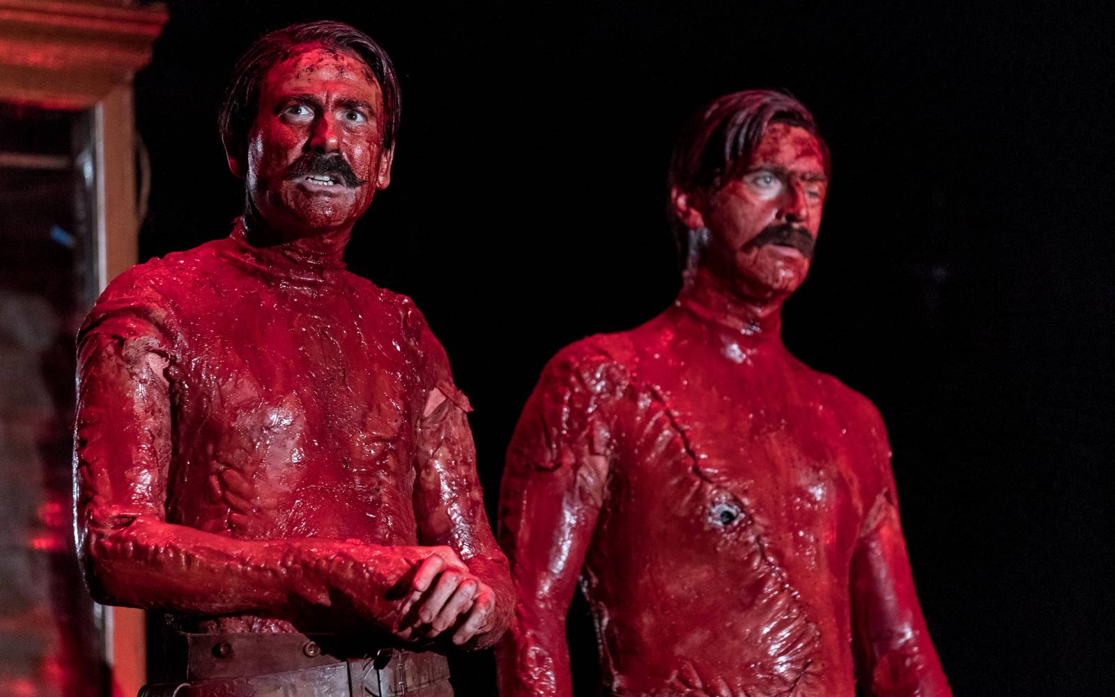 Two topless men, covered all over in blood. Their bodies are riddled with scars