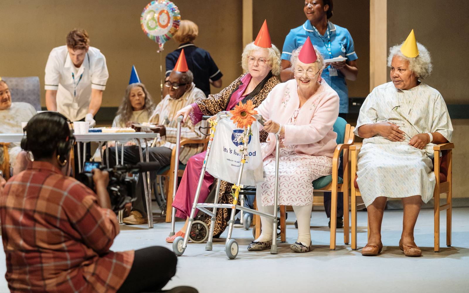 Elderly patients sit on chairs in a hospital. They are wearing paper party hats. Medical staff attend to them. In the foreground, with her back to the camera, a camerawoman films the scene