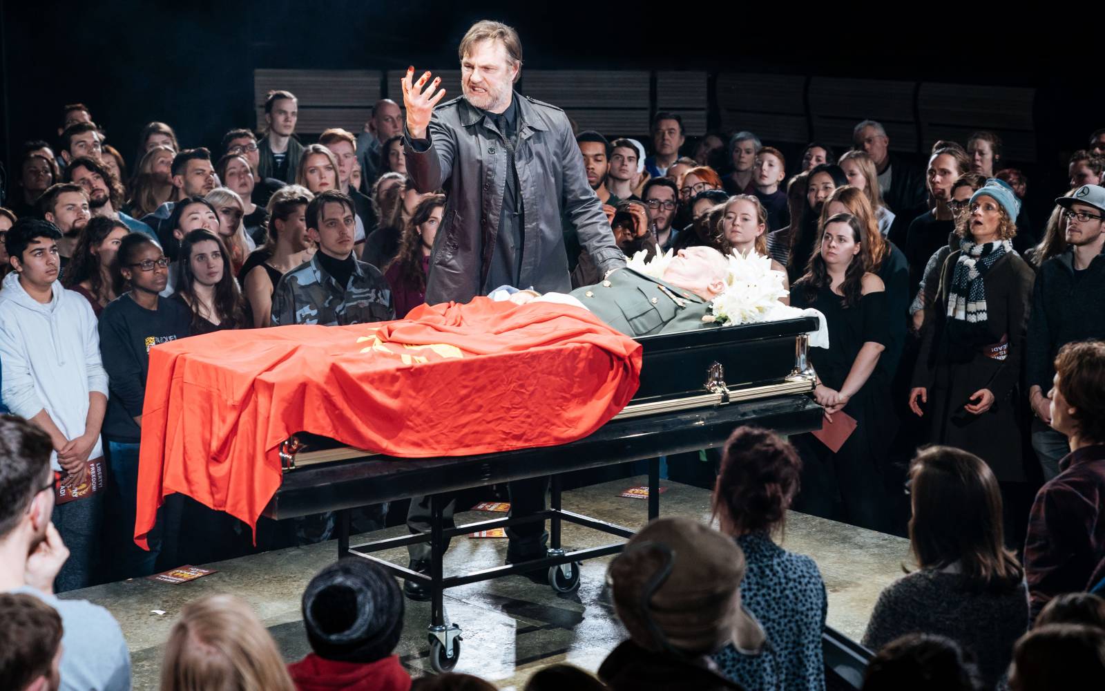 A man stares at his hand with fury. He stands over a body in a coffin, draped in a red flag, surrounded by a crowd