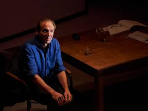 Taken from a higher angle looking down on the stage. We see a man (Ralph Fiennes) sitting on a chair next to a large wooden desk. On the desk are open books, glasses, glasses case, a glass jug. Ralph is wearing a blue shirt with the long sleeves folded up to his elbow. His hands are together in his lap and he looks lost.