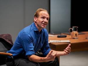 A man (Ralph Fiennes) sat in a chair next to a large wooden desk. The stage is well lit. Ralph is leaning forward with his elbows resting on his knees and his hands out as if explaining something. He's in a blue shirt with the sleeves rolled up to the elbows.