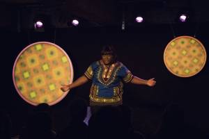 Taken from the back of the room, we can see silhouettes of the audience heads. A dark stage but with 2 illuminated circles either side of Yolanda. They both have orange, yellow and green prints on them but no words. In the middle stands Yolanda in a blue, yellow and pink dashiki top. Her fringe is down across her forehead and her arms are held out by her sides as if asking a question.