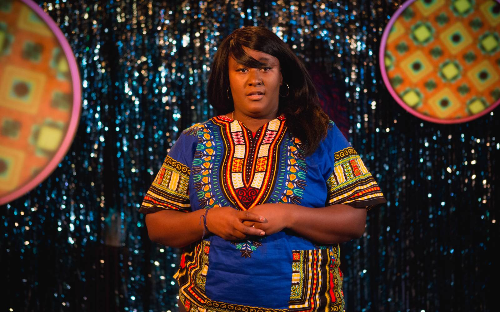 A sparkly blue tassled backdrop. A woman (Yolanda Mercy) is stood in the centre, wearing a dashiki top with bold blue, reds and yellows. Her hands are entwined in front of her and she's looking out into the audience. She's wearing large hoops in her ears and her hair is long, straight with a side fringe sweeping over her face.