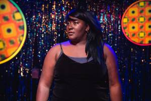 A blue sparkly tassle background to the set. Yolanda Mercy stands in front wearing a strappy black velvet top. Her straight dark hair is pushed over one shoulder, and she looks out in to the audience the other way.
