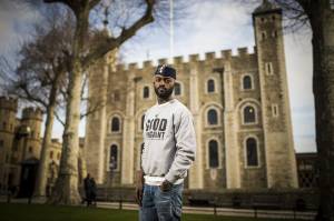 Inua Ellams, a man in his 30s, is stood in front of an out of focus Tower of London. He's wearing blue jeans and a grey sweatshirt that has black text on it reading 'the good immigrant'. He has one hand in his pocket and you can see a black watch on that wrist. He's wearing a black Zanna Bukar Nigerian cap with buttons and pins on one side at the front.