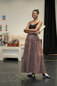 Photo of Racheal Ofori. She's wearing a black strappy top, long rehearsal skirt and black small heels. Her arms are folded and she's holding a scroll in one of her hands, she's laughing with a huge grin.