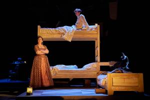 Lit on the stage is a wooden bunk bed and a small wooden bed which has a young boy sleeping in it. Samuel Blenkin is sat upright on the top bunk in a night shirt. Pandora Colin is leaning against the bunk bed frame with her arms crossed and looking off into the distance.
