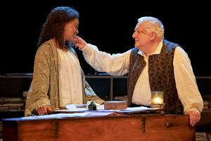 Racheal Ofori and Simon Russell Beale are stood behind the harpsicord. They're looking at each other and his hand is touching her cheek.