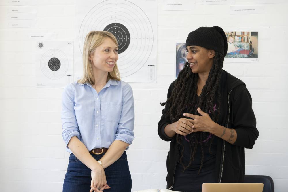 Polly Findlay (blonde bob, pale blue shirt) and Suzan-Lori Parks (long twisted braids, black beanie and hoodie) stand in the rehearsal room. They are smiling in conversation with each other. Behind them are mood board images including a target from a shooting range.