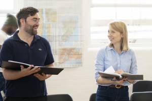 Rehearsal photo of James Corrigan (silver framed glasses, rugged bear and dark blue polo shirt) and Polly Findlay (blonde bob and blue shirt). They are both holding scripts and are looking at each other laughing.