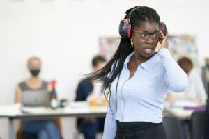 Rehearsal photo of Faith Omole (black braids tied up, pale blue collared top with a zip down the front). She is wearing protective eye glasses and noise defenders over her head. Her hand is held up to the side of her face and she's captured mid turn, so her braids are mid swish.