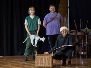 Heather Forster (blonde hair tied back, white tshirt with green sweater vest) holds origami style puppet of a cat daemon. Samuel Creasey (blonde hair, purple t-shirt), stand behind Ella Dacres (cream beanie, black jumper) who crouches next to a wicker basket.