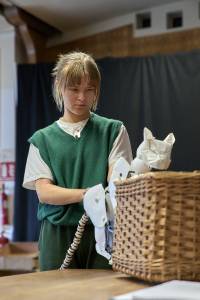 Heather Forster (blonde hair tied back, white tshirt with green sweater vest) holds origami style puppet of a cat daemon peering into a wicker basket.