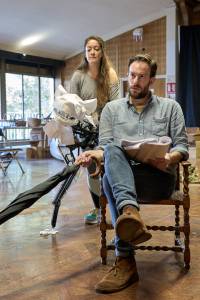 Pip Carter, (brown hair, brown beard) sits on a chair, he is holding an umbrella. Julie Atherton (long brown hair, grey top), holding a large origami style hyena puppet.