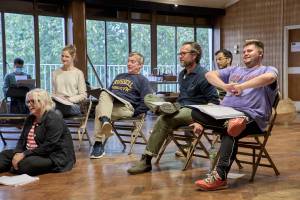 Naomi Frederick (blonde hair tied back, white jumper), Nick Sampson (short grey hair, navy jumper), John Light (brown hair, glasses, navy shirt), Sid Sagar (black hair, glasses, green shirt), Samuel Creasey (short blonde hair, purple t-shirt). Seated on chairs. Holly Atkins (white short hair, glasses, denim jacket, red and white striped top), sits on the floor.