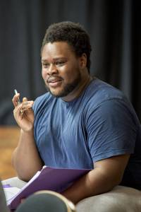 Tomi Ogbaro (short black hair, black beard, blue t-shirt), sits in rehearsals. He is holding a script and open.