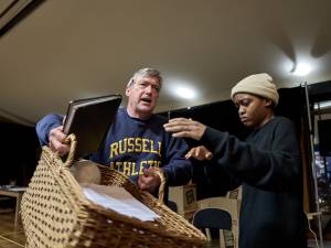 Nick Sampson (grey hair, navy jumper) holds a wicker basket as Ella Dacres (black hair, cream beanie, black jumper) stands next to him and reaches towards the basket.