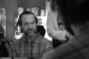 John Light (Dark hair swept back and tucked behind his ears, checked shirt and a beard) sits in his dressing room, smiling.