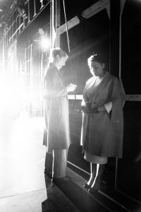 Naomi Frederick (blonde hair tied back and a long coat) stands back stage waiting to go on with Ayesha Dharker (hair tied back, long coat and heels). The light from on stage illuminates the two from behind and the light flares across the image.