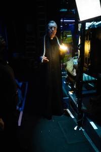 Nick Sampson stands backstage with a snake puppet draped over his shoulders. A light in the puppet illuminates his face with a warm yellow light.