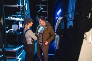 Olivia Le Anderson (Blonde hair in bunches, a beanie and dungarees) and Samuel Creasey (fair hair, patterned sweater vest and a green coat) stand backstage whispering. In the background various props can be seen.