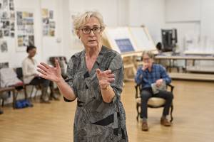 Helen Schlesinger (short white hair, glasses and grey patterned dress) in rehearsals. She stands and looks off camera with her hands splayed out as though explaining something.