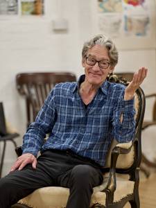 Guy Paul (grey wavy hair, glasses and a blue checked shirt) sits in an old wooden chair leaning on one armrest gesturing with a smile on his face.