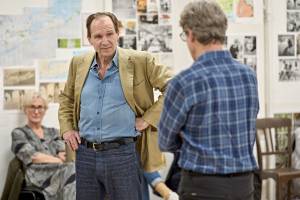 Ralph Fiennes (brown hair, blue collared shirt and a beige blazer) stands facing the camera with a fisted hand on his hip. He looks at Guy Paul (grey hair, glasses and blue checked shirt) who stands facing away from the camera with arms folded.