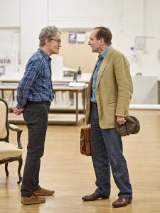 Guy Paul (grey hair, glasses, blue checked shirt) stands hands behind his back opposite Ralph Fiennes (brown hair, blue collared shirt, beige blazer) who holds a brown satchel in one hand and clutches a flat cap in the other.