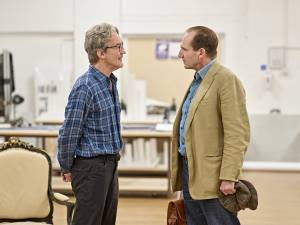 Guy Paul (grey hair, glasses, blue checked shirt) stands hands behind his back and smiling opposite Ralph Fiennes (brown hair, blue collared shirt, beige blazer) who holds a brown satchel in one hand and clutches a flat cap in the other.