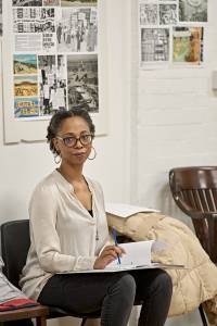 Alana Maria (black hair tied back, glasses, hoop earrings and cream shirt) sits with a script and pen. Behind her are images of protests on a moodboard in the rehearsal room.