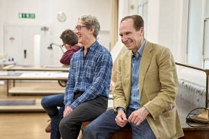 Guy Paul (grey hair, glasses and blue checked shirt) sits with Ralph Fiennes (brown hair, blue shirt and beige blazer) they are laughing and smiling.