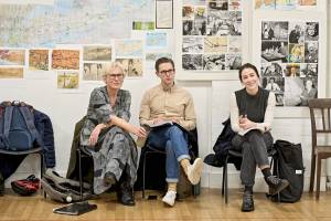 Helen Schlesinger (short white hair, glasses and grey patterned dress), Samuel Barnett (brown hair, glasses, beige shirt, blue jeans), Siobhán Cullen (long dark hair tied back, grey knitted vest over a longsleeved white top) all sit on chairs in rehearsal with scripts on their laps. They look focused as Siobhán smiles.