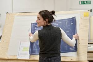 Siobhán Cullen (long dark hair tied back, grey knitted vest over a long-sleeved white top) faces away from the camera and holds a blueprint map on a large wooden board.