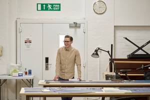 Samuel Barnett (brown hair, glasses, beige shirt) stands smiling, he is leaning on a table covered in blueprints.