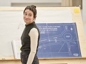 Siobhán Cullen (long dark hair tied back, grey knitted vest over a long-sleeved white top) stands in front of a wooden board holding a blueprint. She smirks and looks over her shoulder.