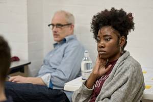 Dani Mosely (black hair, hoop earrings and a grey cardigan over a burgundy tshirt) sits in rehearsals, behind her is Ian Kirkby. They both look focused.