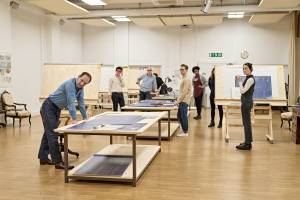 The cast of Straight Line Crazy stand looking focused, there are wooden tables and boards with blueprints.