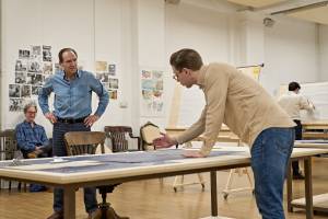 Ralph Fiennes (brown hair, blue collared shirt and a beige blazer) stands hands on hips. He looks at Samuel Barnett (brown hair, glasses, beige shirt), who leans over a table and gestures to the blueprints in front of him.