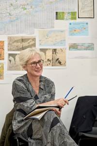 Helen Schlesinger (short white hair, glasses and grey patterned dress) in rehearsals. She sits with her script in her lap smiling.